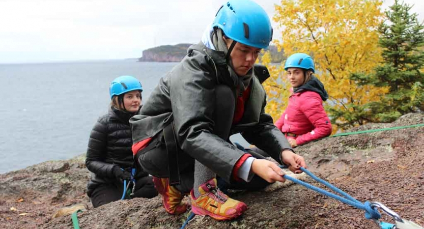 A person wearing safety gear pulls on ropes secured to a rock. There are two people wearing helmets in the background. They all appear to be on a cliff above a body o f water. 